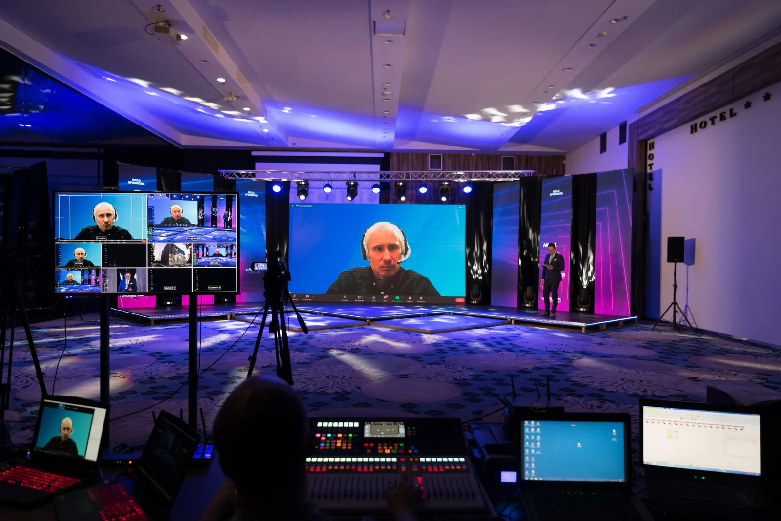 Hybrid conference production setup with cameras, laptops, monitors, and AV gear inside a convention center session room