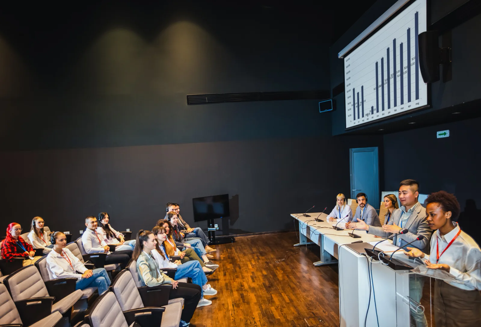 Public hearing room with presentation displays, microphones, staff laptops, and attendees seated for a government meeting
