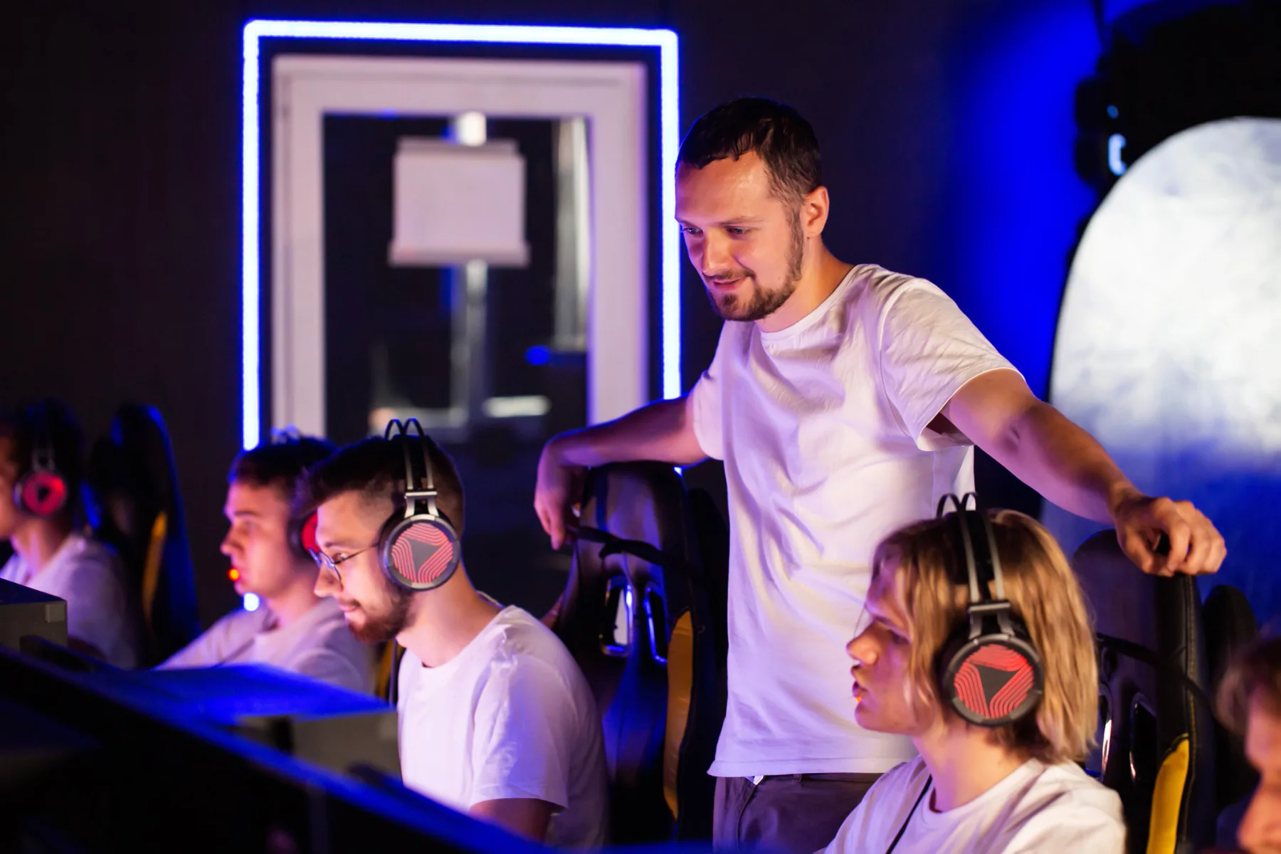 Esports team practice room with multiple gaming setups, coaches reviewing screens, and bootcamp training environment