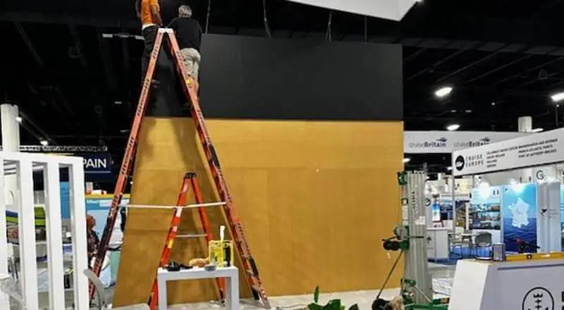 A blank wall at a trade show with work equipment around it. A man stands on top of a ladder working on some wiring.